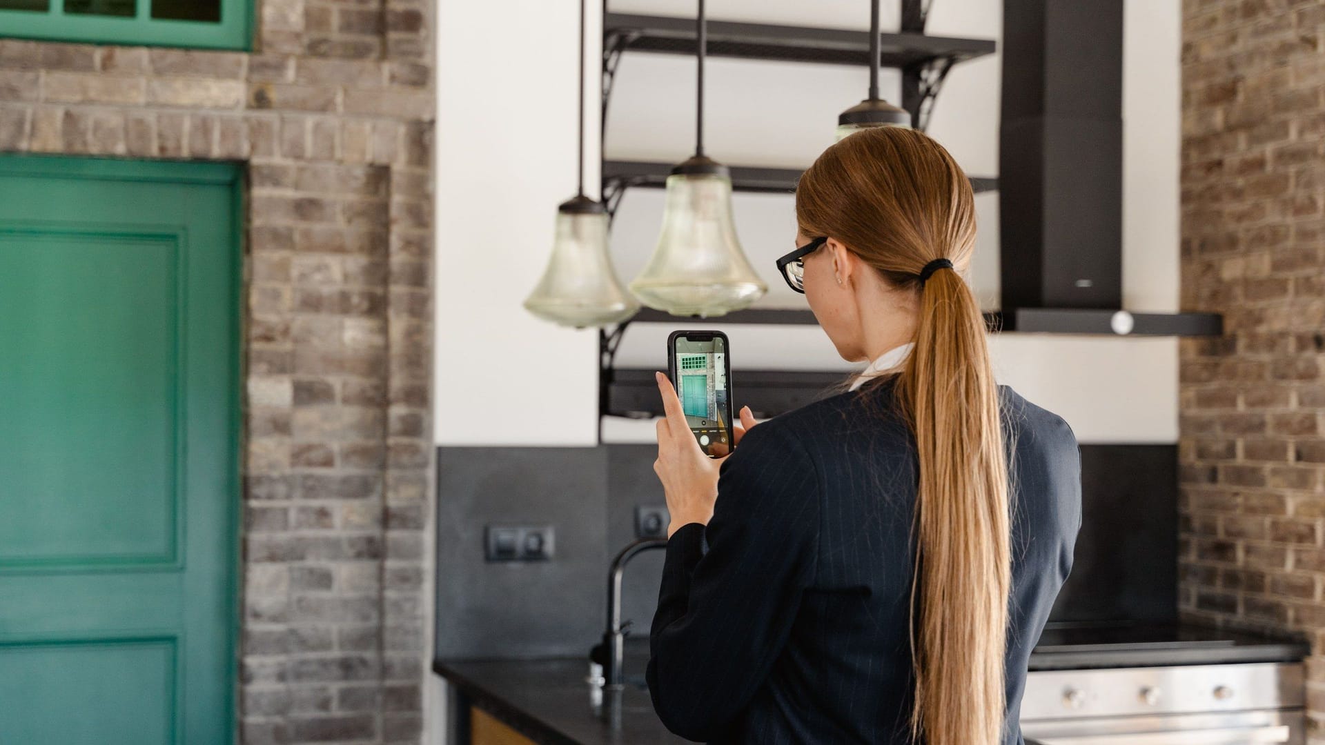 A woman photographs a kitchen with her phone, with glass pendant lights, a brick wall and a green door in view.