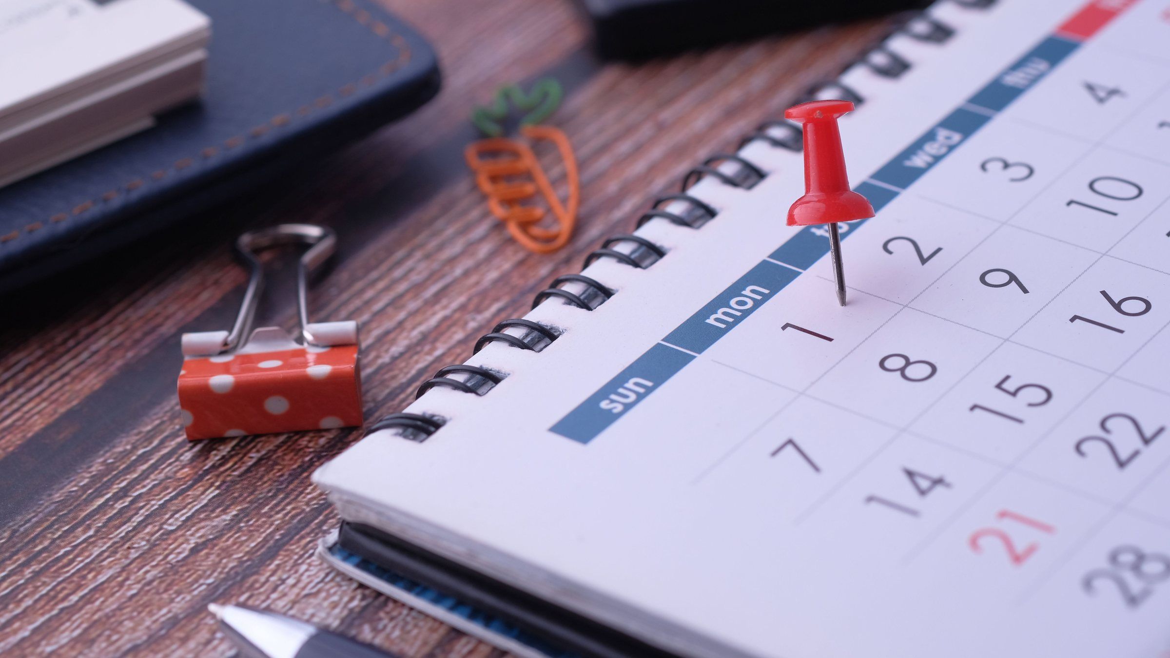 A red push pin marks a single date on a spiral-bound desk calendar next to a binder clip and a pen.