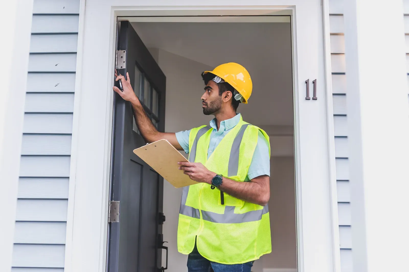 A man in a hard hat and safety vest holds a clipboard while he checks the frame of a house's front door.